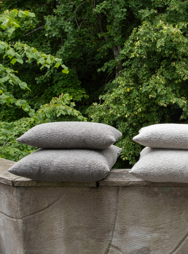1K_Hynne-outdoor-Pebble-Mix_23 Stacks of gray Hynne outdoor cushions arranged on a low stone wall, with trees and foliage in the background.