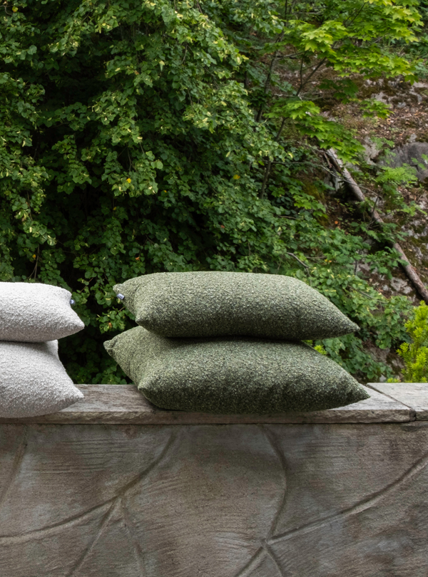1K_Hynne-outdoor-Pebble-Mix_24 Stacks of gray and green Hynne outdoor cushions arranged on a low stone wall, with trees and foliage in the background.