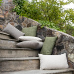 Outdoor scene featuring Hynne cushions in cream, gray, and green, placed on stone steps leading up to a stone wall with greenery.