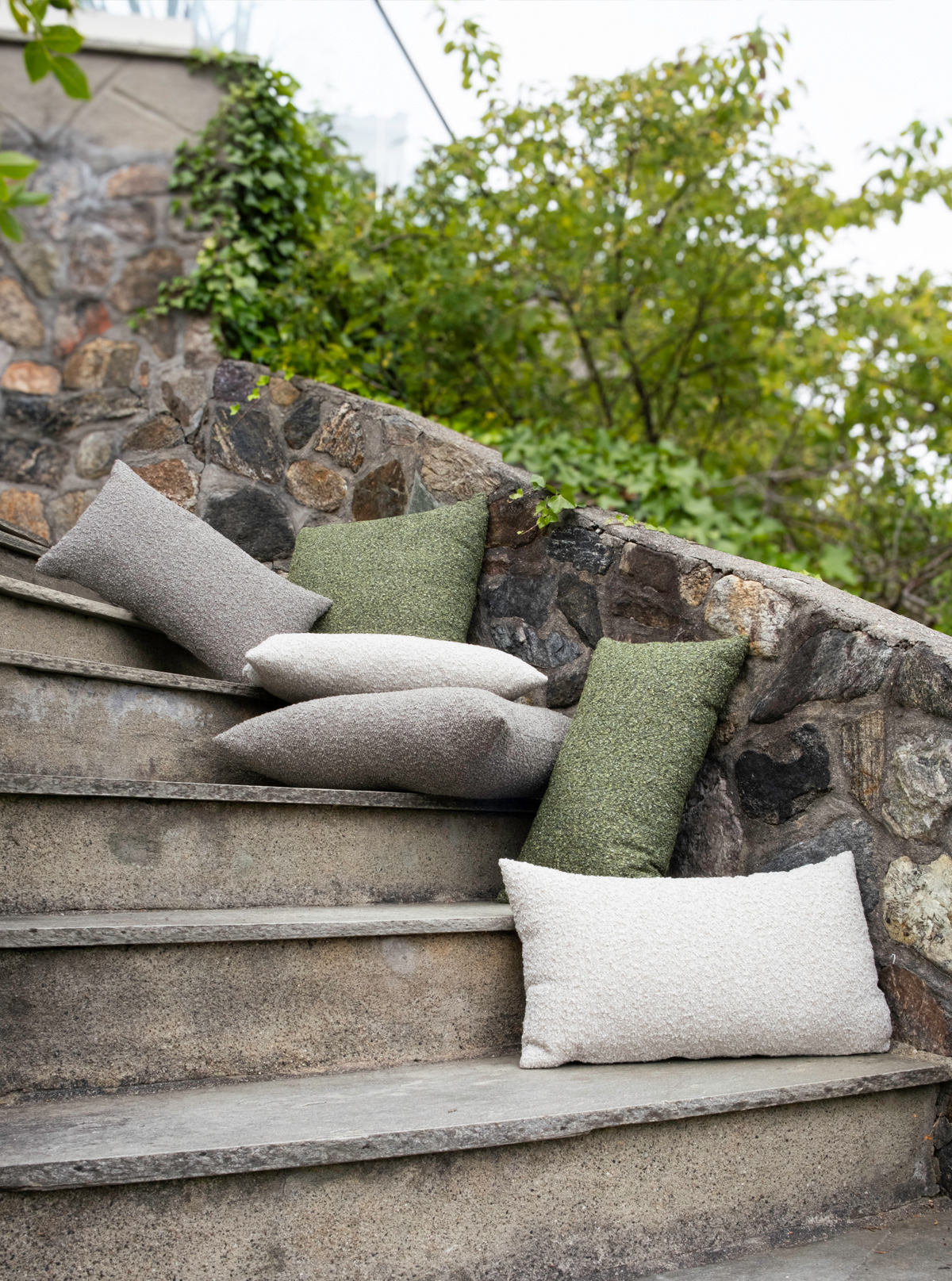 2K2H_Hynne-outdoor-Pebble-Mix_20 Outdoor scene featuring Hynne cushions in cream, gray, and green, placed on stone steps leading up to a stone wall with greenery.