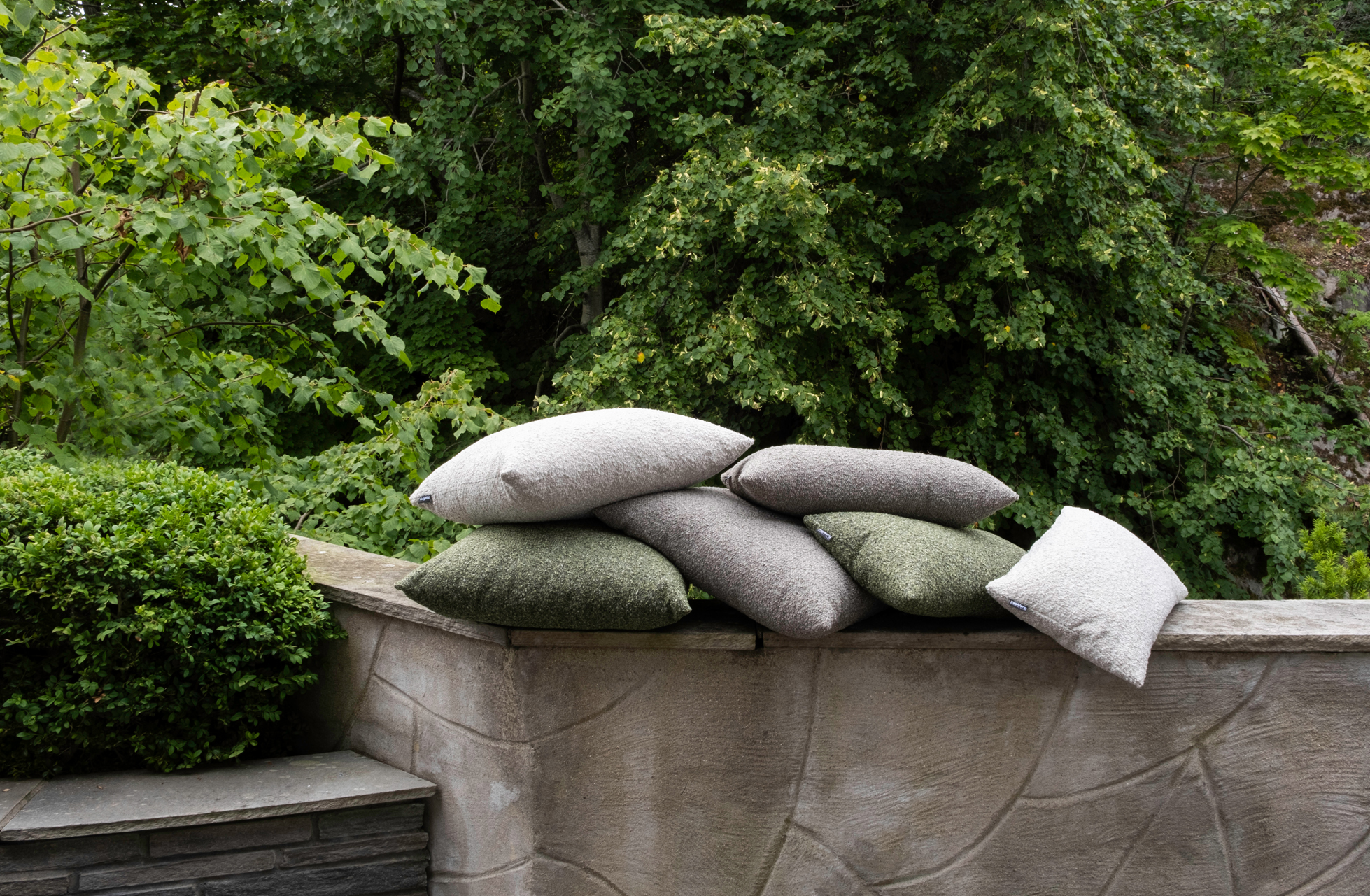 3K_Hynne-outdoor-Pebble-Mix_21 Several Hynne outdoor cushions in gray, light gray, and green, stacked on a stone wall with a leafy backdrop.