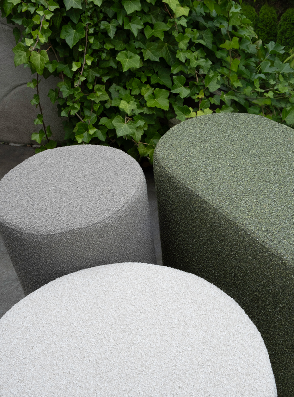 Three Mingle poufs in light gray, dark gray, and green on a stone patio with greenery in the background.