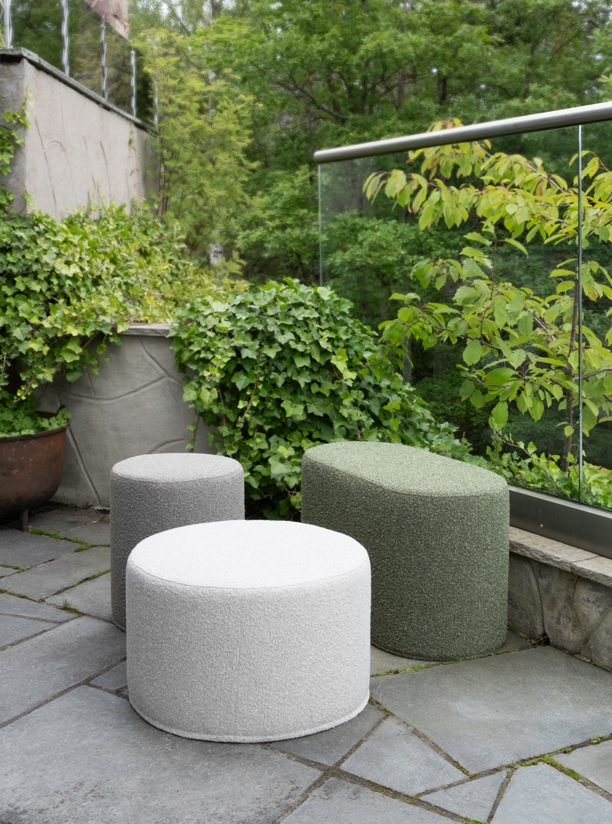 Three Mingle outdoor poufs in varying shapes and colors (cream, gray, and green) arranged on a patio with green plants and a glass railing in the background.