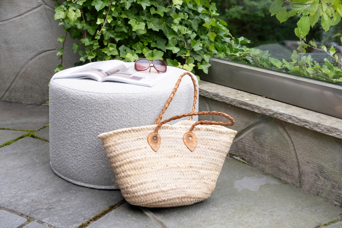 Outdoor patio featuring a round, light gray Mingle pouf with an open magazine and sunglasses on top, beside a woven straw bag. Background includes lush green ivy and a stone wall.
