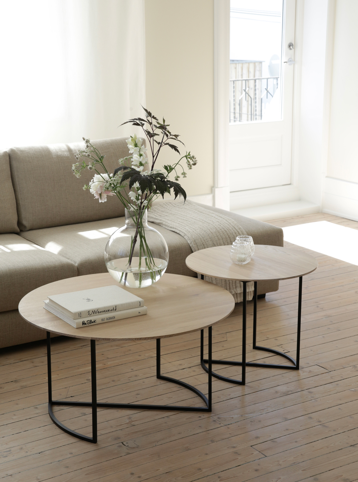 Alt text: "Modern living room with Ljå coffee table set, featuring light wooden tops and black metal legs, complemented by a vase of flowers and books.