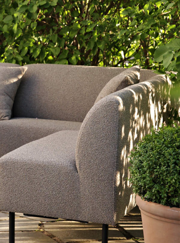 1K_Sofa-Aften-outdoor-62-Pebble-Birch_22 Gray Aften outdoor dining sofa with a small round table on a stone patio, surrounded by greenery and dappled sunlight.