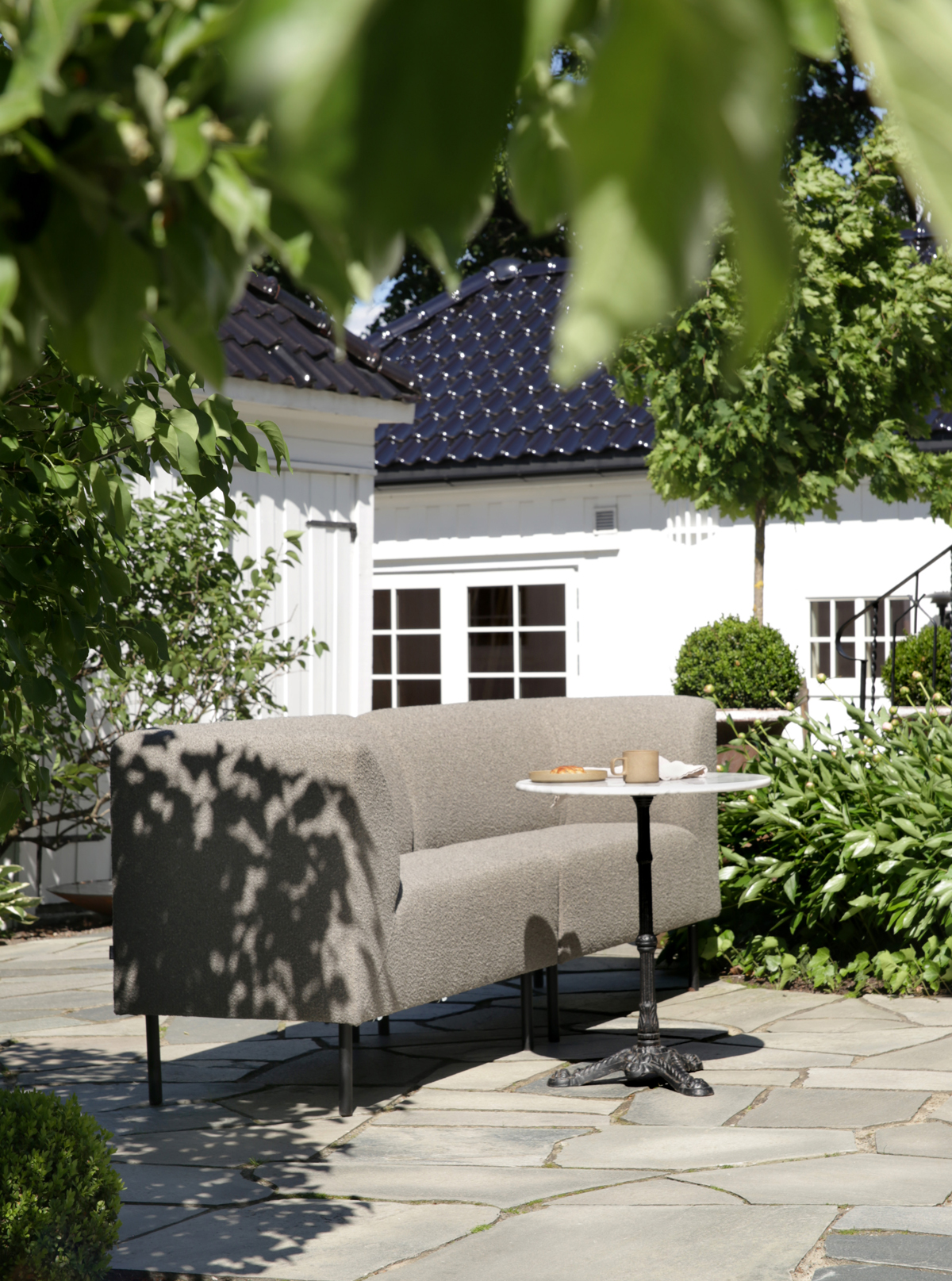 2K2H_Sofa-Aften-outdoor-62-Pepple-Birch_06 Stacks of gray and cream Hynne outdoor cushions arranged on a low stone wall, with trees and foliage in the background.