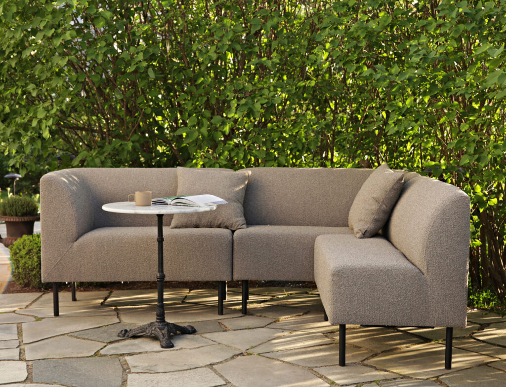 Gray Aften outdoor dining sofa with a small round table on a stone patio, surrounded by greenery and dappled sunlight.