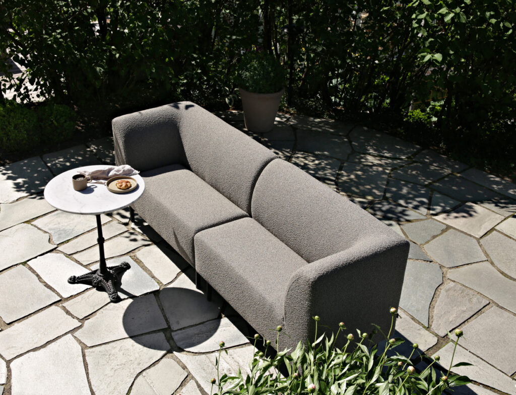 Gray Aften outdoor dining sofa with a small round table on a stone patio, surrounded by greenery and dappled sunlight.