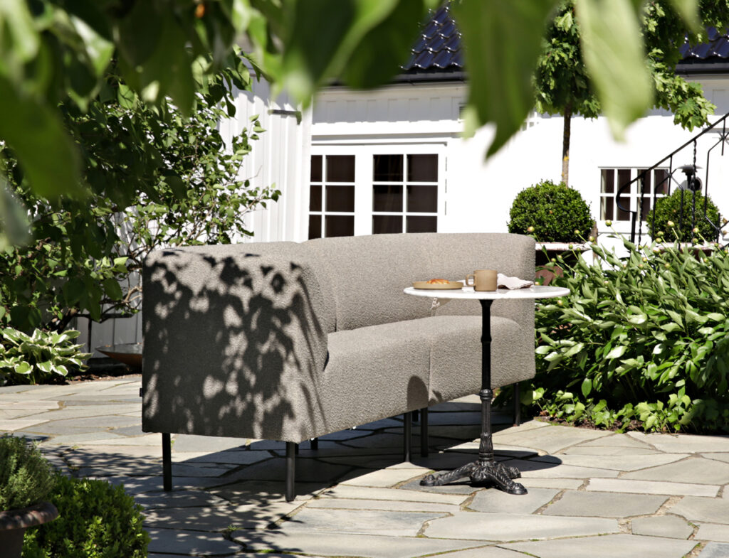 Gray Aften outdoor dining sofa with a small round table on a stone patio, surrounded by greenery and dappled sunlight.
