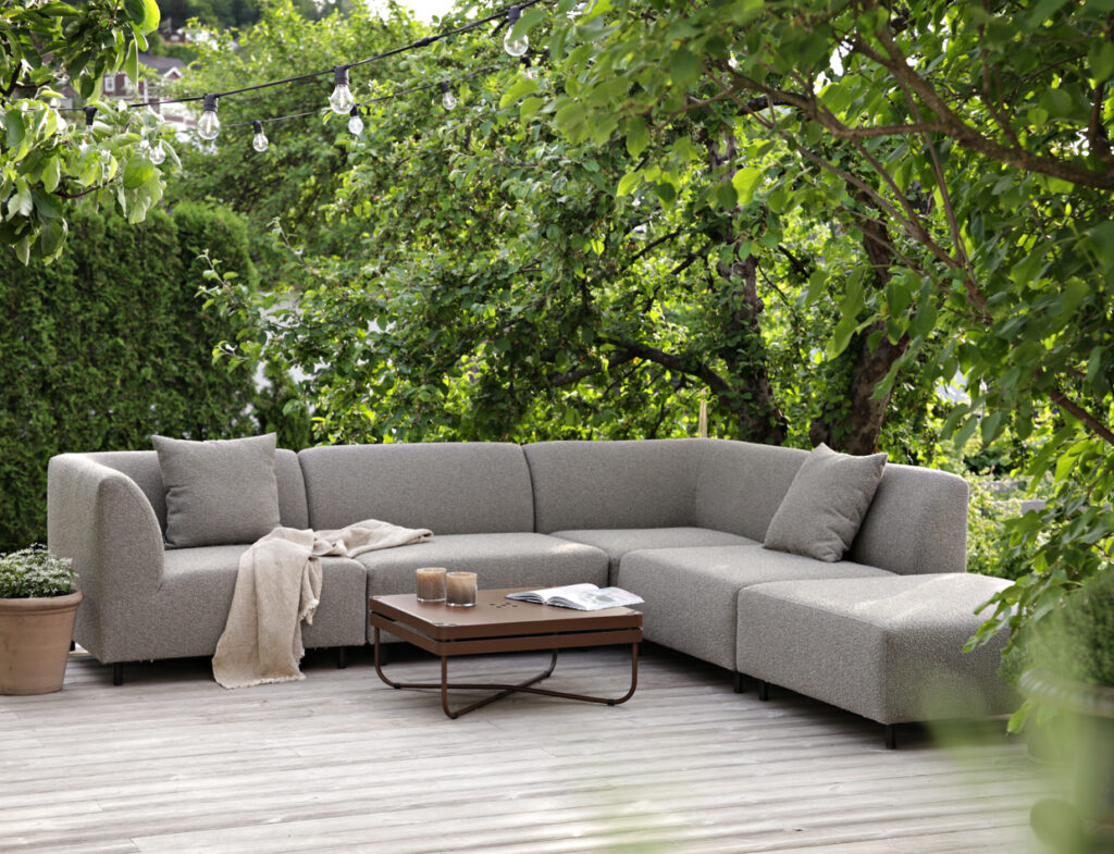 A light gray Aften modular outdoor sofa and a coffee table on a wooden deck, surrounded by greenery.