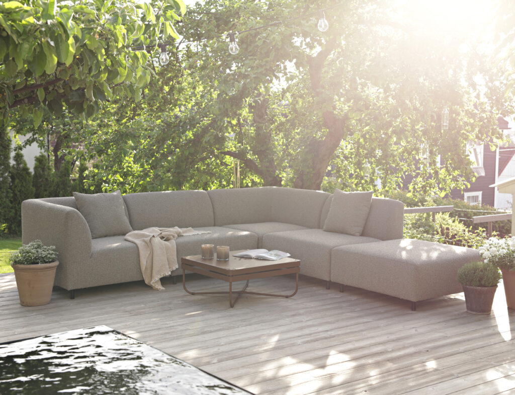 A light gray Aften modular outdoor sofa and a coffee table on a wooden deck, surrounded by greenery and string lights.