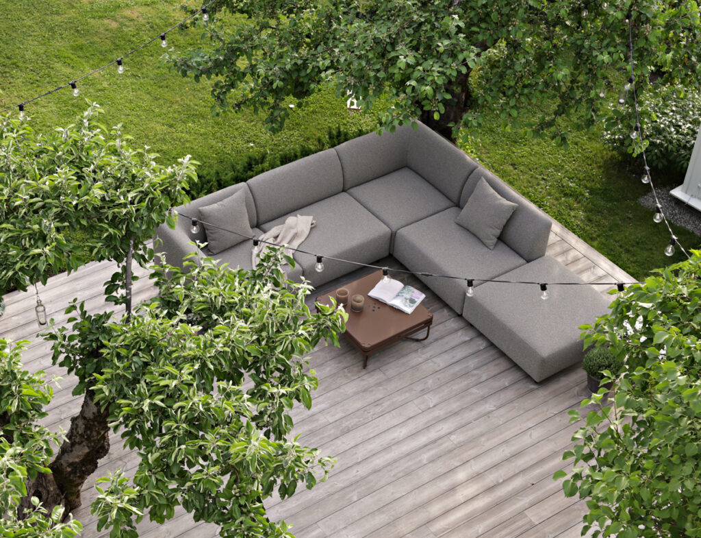 A light gray Aften modular outdoor sofa and a coffee table on a wooden deck, surrounded by greenery.