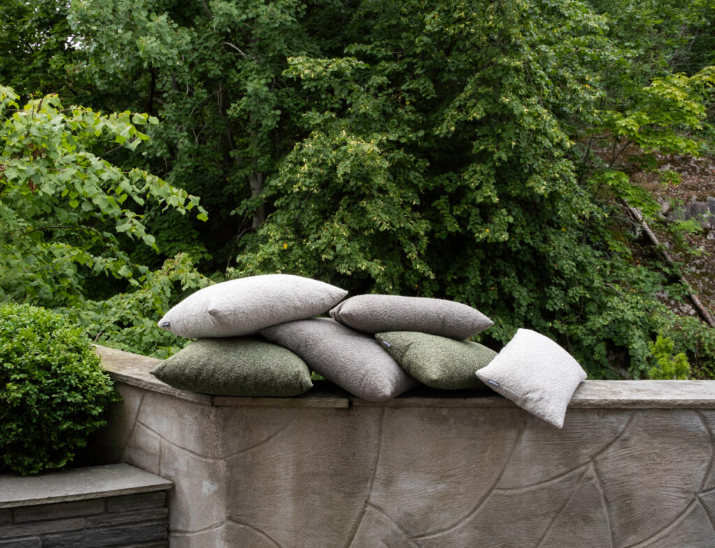Several Hynne outdoor cushions in gray, light gray, and green, stacked on a stone wall with a leafy backdrop.
