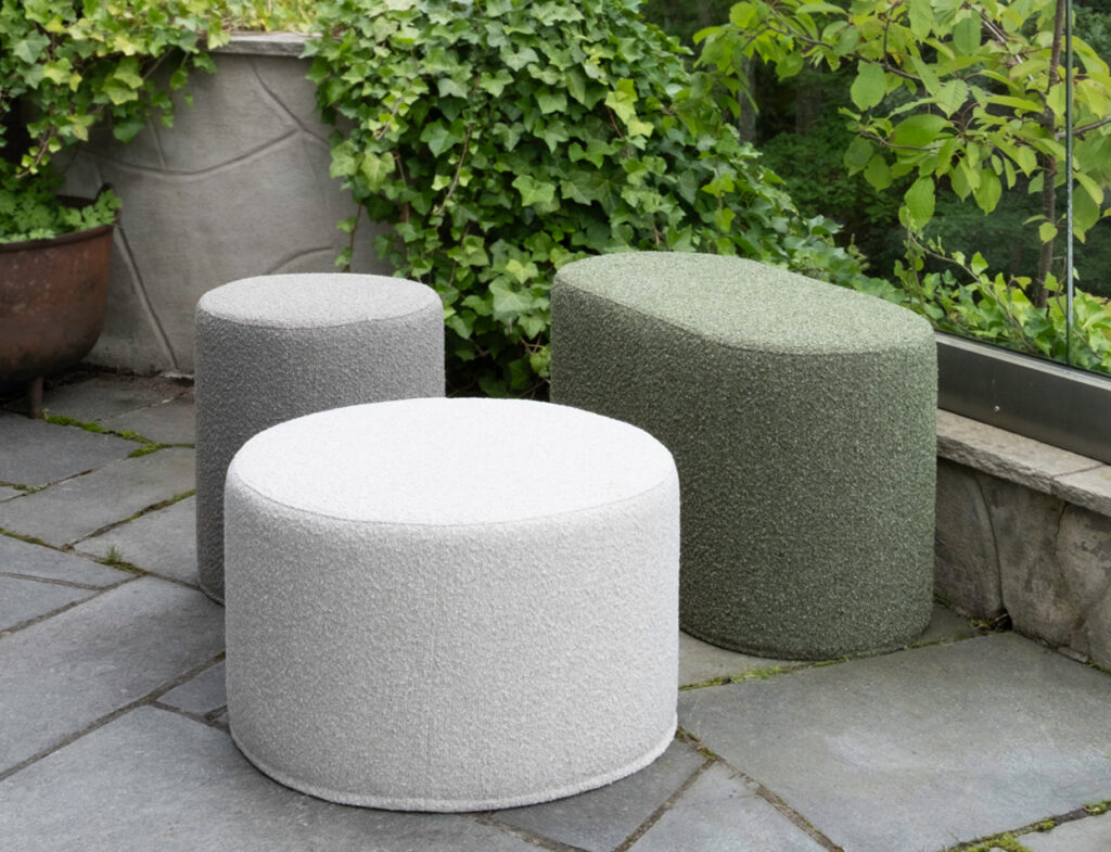 Three Mingle poufs in light gray, dark gray, and green on a stone patio with greenery in the background.