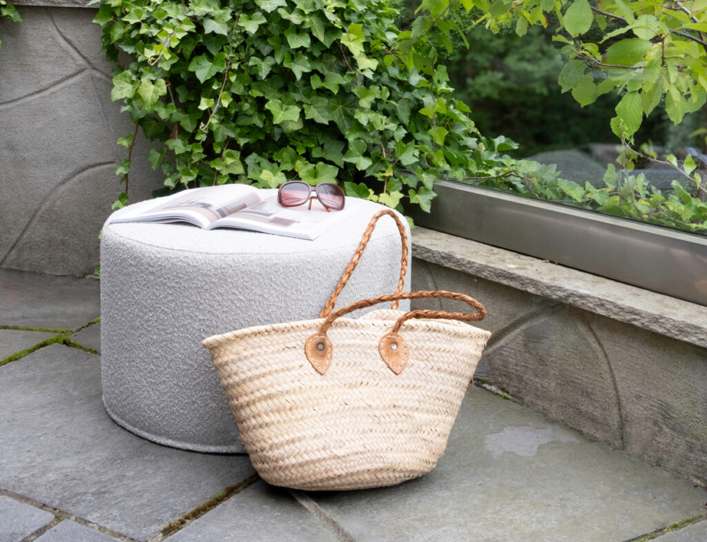 Outdoor patio featuring a round, light gray Mingle pouf with an open magazine and sunglasses on top, beside a woven straw bag. Background includes lush green ivy and a stone wall.