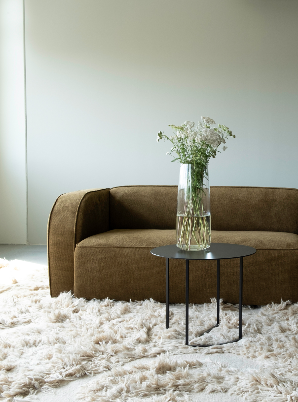 2K2H_NySofa-Yme_03 A brown Sva module sofa sits on a cream-colored shaggy rug, accompanied by a small black side table with a vase of white flowers.